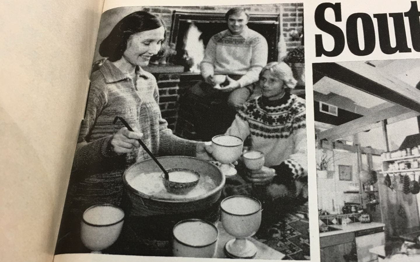 A white woman dressed in 1970s fashion services soup to two white men. All are smiling. 