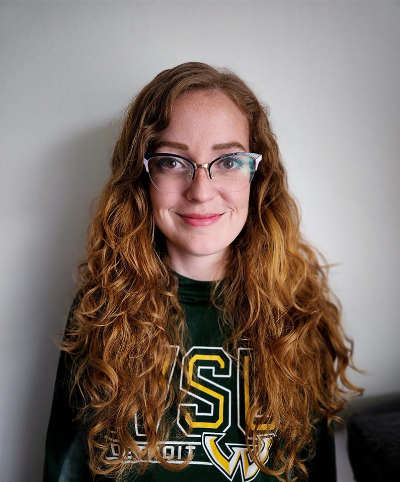 Headshot of Carly Slank. She has long red hair, and is wearing dark top-framed glasses and a Wayne State University sweatshirt.