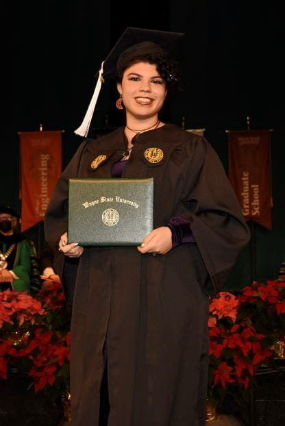 Three-quarter-length photograph of Rebecca Phoenix. She has black hair in an updo and is wearing a cap and gown, holding her diploma.