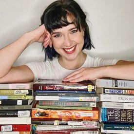 Headshot of Jocie Osika. She has chin-length black hair with bangs, is wearing a white t-shirt, and is leaning on a pile of books in front of her.