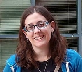 Headshot of Aimee Shulman. She has shoulder-length brown hair and is wearing blue-framed glasses, a black shirt, and a blue sweatshirt.